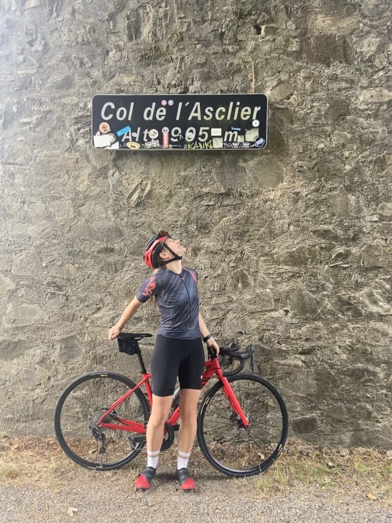 Judith vor ihrem Fahrrad. Sie guckt auf ein Schild, auf dem "Col de l'Asclier" steht.